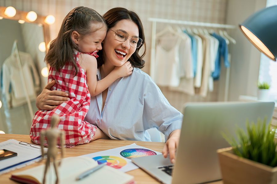 Business Insurance - Smiling Small Business Owner Working On Her Laptop While Hugging Her Daughter